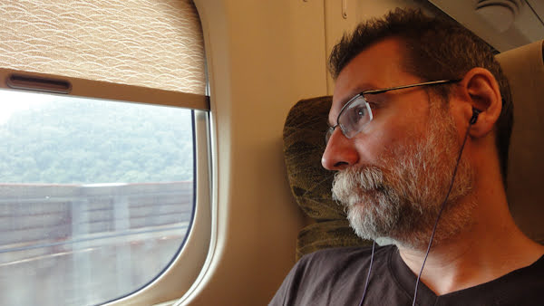 Man with short beard and hair wearing glasses looks out a train window